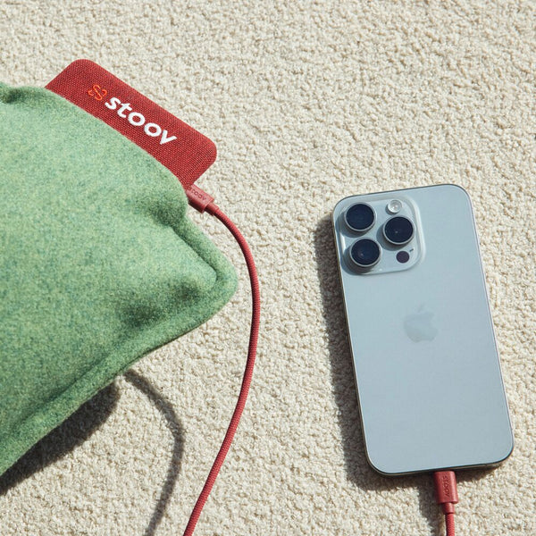Ein Stoov-Heizkissen und ein silbernes iPhone mit roten Ladekabeln auf einem beigen Teppich.