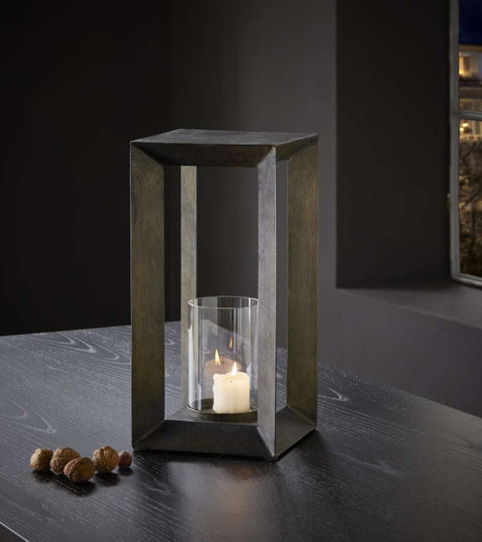 Eine brennende Kerze in einem Glashalter befindet sich in einem rechteckigen Metallrahmen auf einem dunklen Tisch in der Nähe eines Fensters.