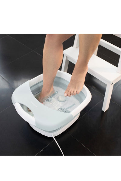 Eine Person legt ihre Füße in ein weißes und hellblaues, mit Wasser gefülltes elektrisches Fußsprudelbad, das auf einem schwarzen Kachelboden neben einem weißen Stuhl steht.