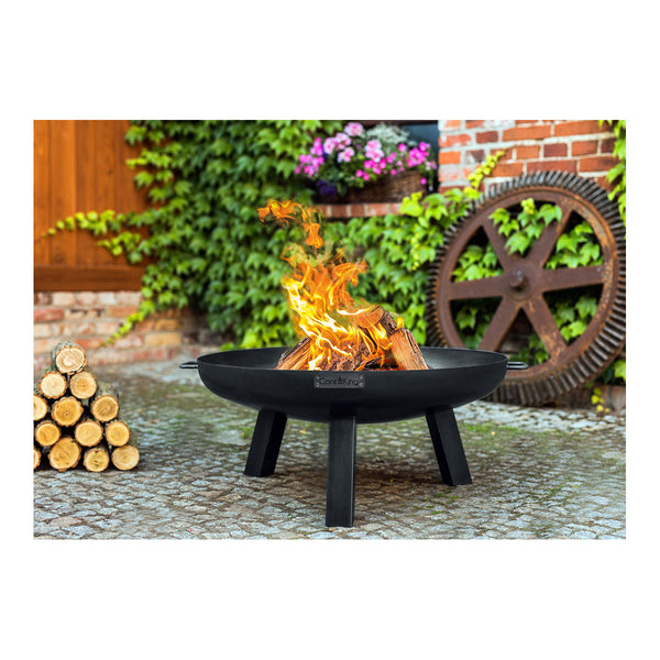 Eine schwarze Feuerstelle mit einem brennenden Feuer, gestapelten Holzscheiten, Blumen und einem großen rustikalen Zahnrad im Hintergrund.