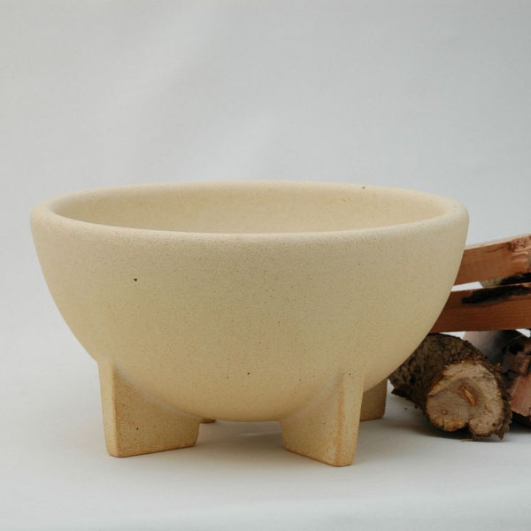 Eine beigefarbene, dreibeinige Keramikschale steht neben gestapelten Brennholzstücken auf einem schlichten Hintergrund.