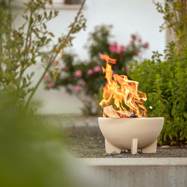 Eine kleine, weiße, moderne Feuerstelle mit brennenden Flammen steht im Freien neben grünen Büschen und rosa Blumen.