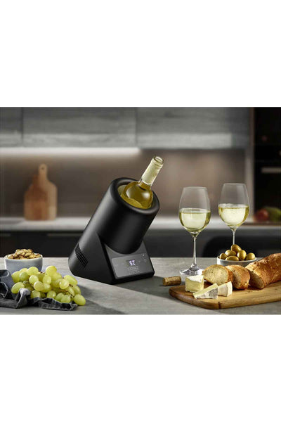 Ein Weinkühler mit einer Flasche Weißwein steht auf einem Küchentisch neben zwei gefüllten Gläsern, Brot, Trauben, Nüssen und Oliven.