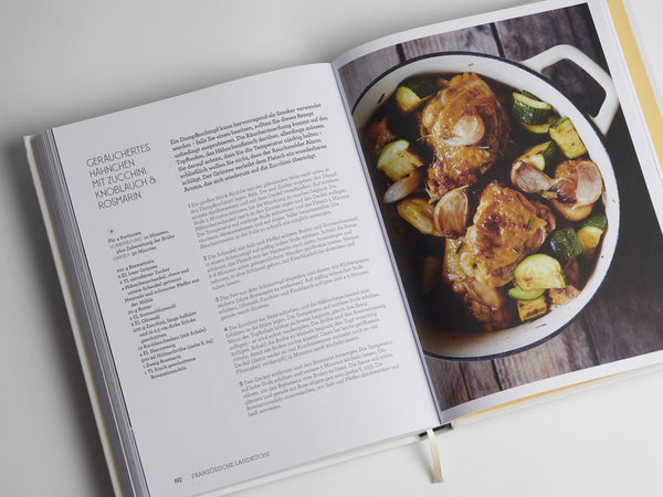 Geöffnetes Kochbuch mit einem Hähnchenrezept und einem Bild von gekochtem Hähnchen und Gemüse in einem weißen Topf.