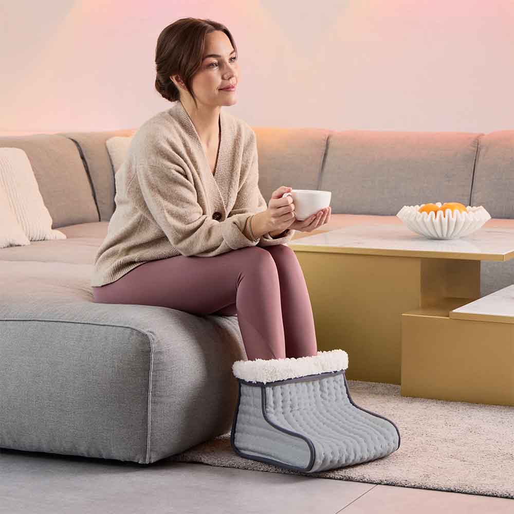 Frau sitzt auf einer Couch, wärmt ihre Füße in einem Fußwärmer und hält eine weiße Tasse.
