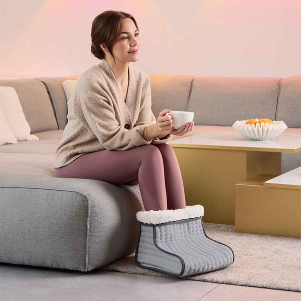 Frau sitzt auf einer Couch, wärmt ihre Füße in einem Fußwärmer und hält eine weiße Tasse.