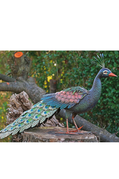 Eine Pfauenskulptur aus Metall mit bunt bemalten Federn steht auf einem Baumstumpf in einem Garten im Freien.