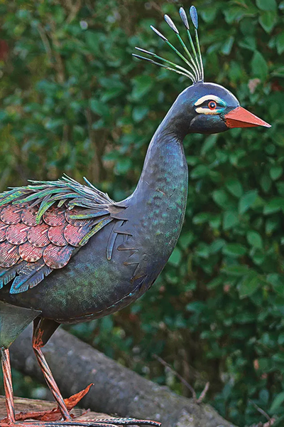 Metallskulptur eines Vogels mit Schopf, grün und bronzefarben schillerndem Gefieder und orangefarbenem Schnabel vor grünem Laub.