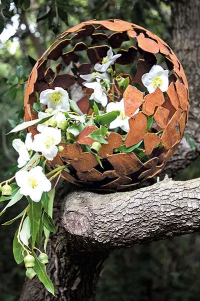 Eine verrostete Metallkugel mit geometrischem Ausschnitt sitzt auf einem Ast, der weiße Blüten und grüne Blätter trägt.
