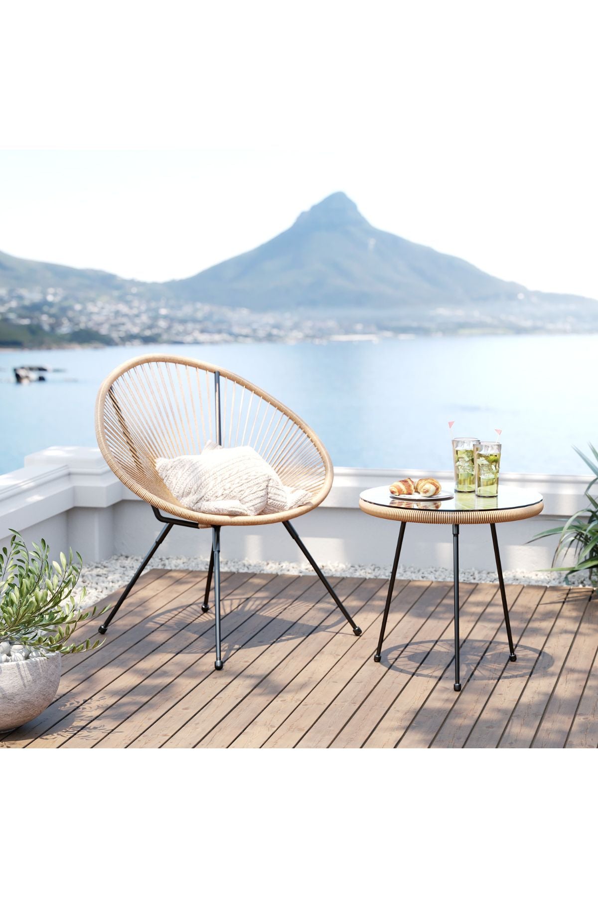 Ein runder Korbsessel mit Kissen und ein kleiner Tisch mit Getränken und Snacks stehen auf einer Holzterrasse mit Blick auf das Wasser und einen fernen Berg.