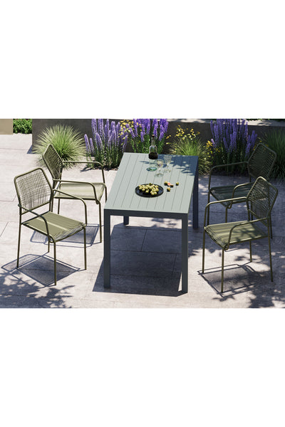 Ein rechteckiger Tisch mit grünlicher Oberfläche ist mit vier Drahtgestellstühlen, Weingläsern und einem Teller mit Snacks gedeckt, umgeben von Terrassenpflanzen und Blumen.