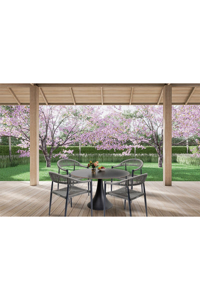 Ein runder Tisch mit vier Stühlen steht auf einer überdachten Holzterrasse mit Blick auf einen Garten mit rosa blühenden Bäumen und grünem Gras.