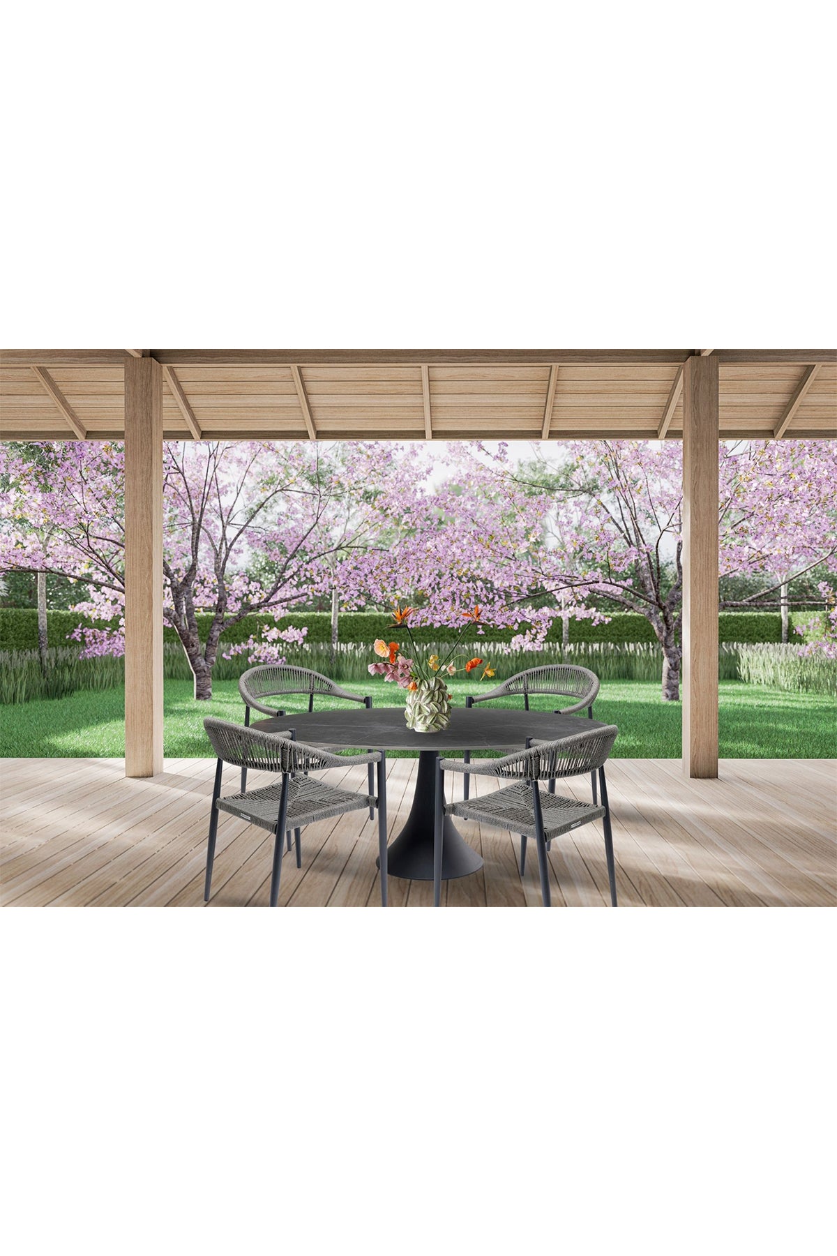 Ein runder Tisch mit vier Stühlen und einer Blumenvase auf einer Holzterrasse mit Blick auf einen Garten mit rosa blühenden Bäumen und grünem Gras.