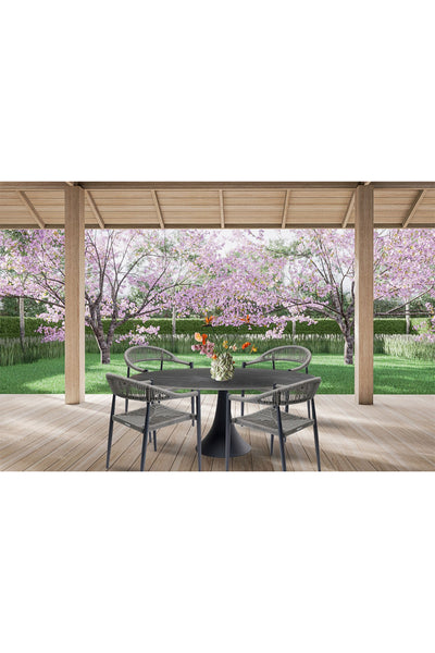 Ein runder Tisch mit vier Stühlen und einer Blumenvase auf einer Holzterrasse mit Blick auf einen Garten mit rosa blühenden Bäumen und grünem Gras.