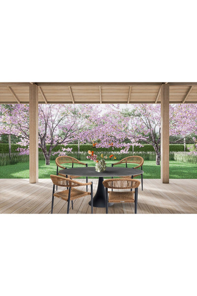 Eine Terrasse mit einem runden Tisch und vier Korbstühlen blickt auf einen Garten mit rosa blühenden Bäumen und grünem Rasen unter einer hölzernen Pergola.