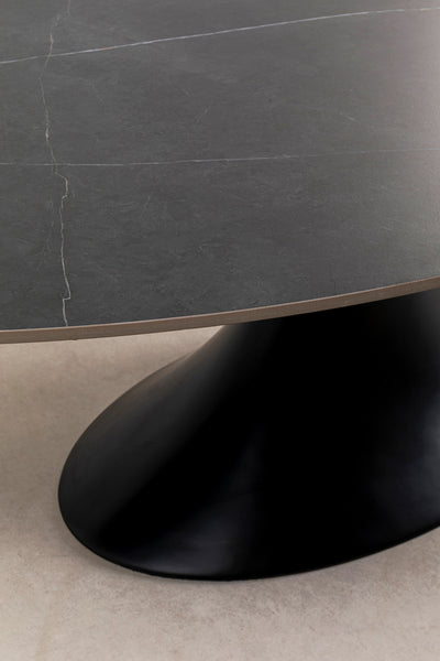 Runder schwarzer Steintisch mit sichtbarer Naht auf der Oberfläche, getragen von einem geschwungenen schwarzen Sockel, der auf einem hellen Boden steht.