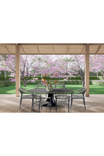 Terrasse mit rundem Tisch, sechs Stühlen und einer Blumenvase mit Blick auf einen Garten mit rosa blühenden Bäumen und grünem Gras.