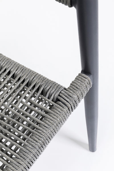 Nahaufnahme eines Stuhls mit geflochtenem Seilsitz und dunkel gefärbten, spitz zulaufenden Metallbeinen vor weißem Hintergrund.