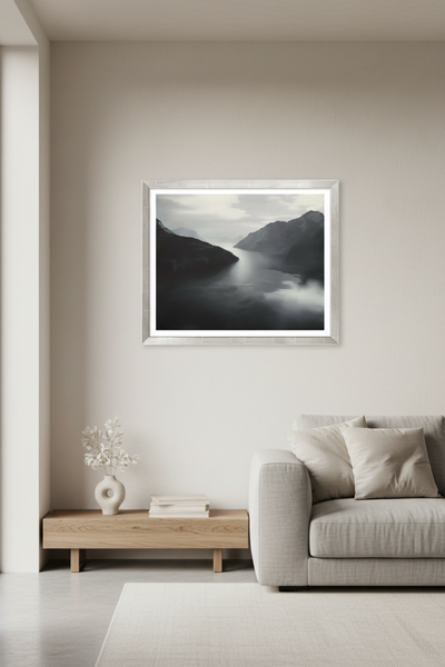 Ein minimalistisches Wohnzimmer mit einem hellgrauen Sofa, einem Couchtisch aus Holz und einem gerahmten Landschaftsfoto mit Bergen und Wasser an einer neutralen Wand.