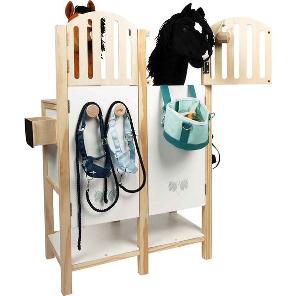 Ein hölzerner Spielzeugpferdestall mit zwei Plüschpferdeköpfen, hängenden Geschirren und einer blaugrünen Tasche, die an den Vorderseiten befestigt ist.