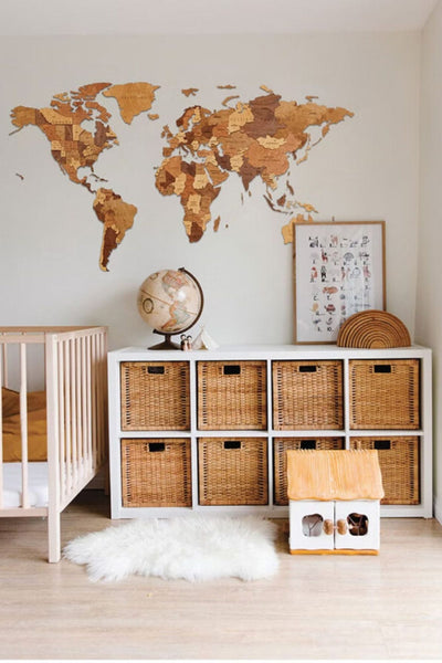 In einem Kinderzimmer stehen ein Kinderbett, ein Weidenkorb, ein Globus, eine Weltkarte, ein gerahmter Druck und ein kleiner Teppich auf einem hellen Holzboden.