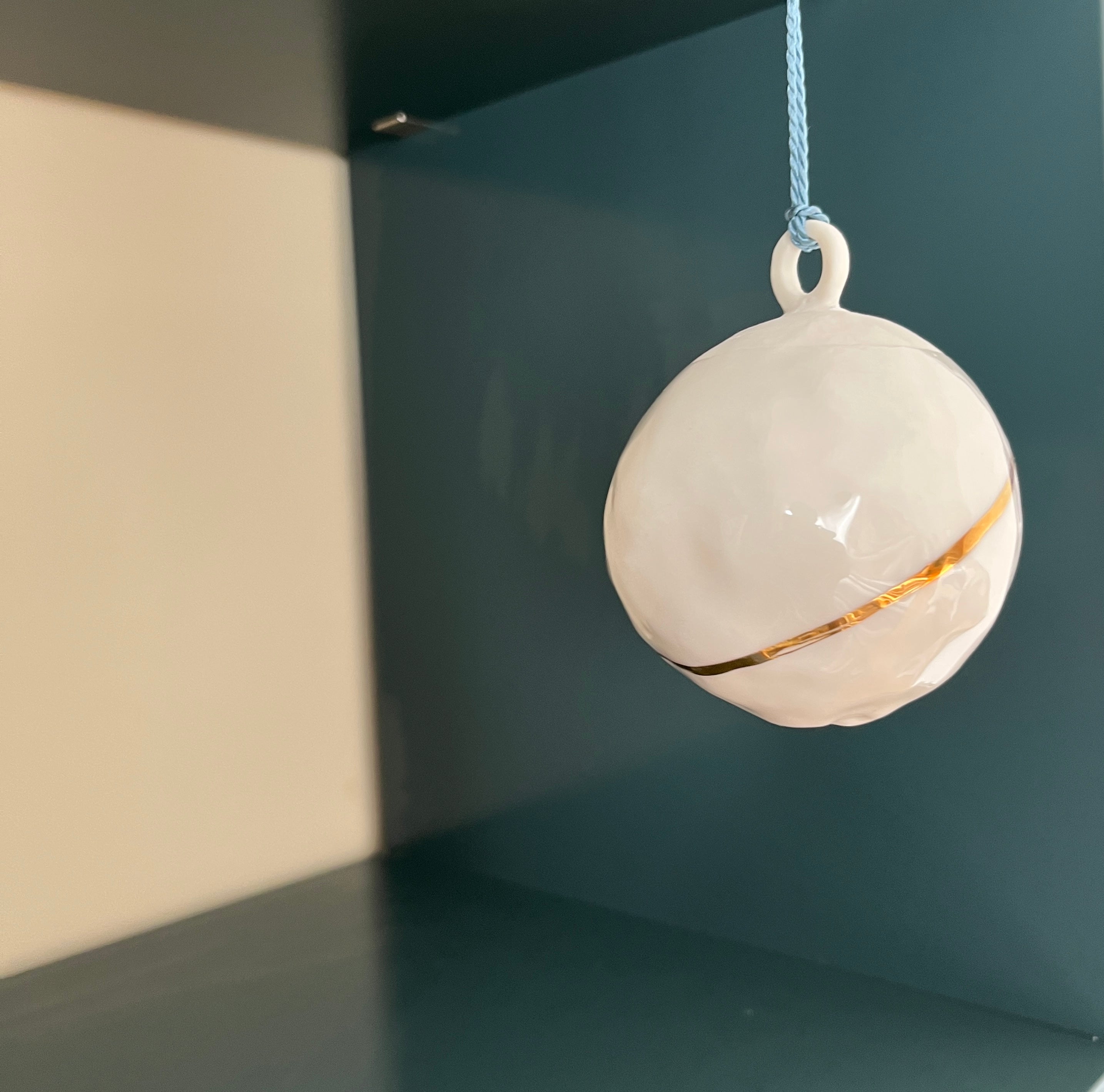 Weißes Ornament mit einem goldenen Streifen, das an einer hellblauen Schnur vor einem dunkelgrünen Hintergrund hängt.
