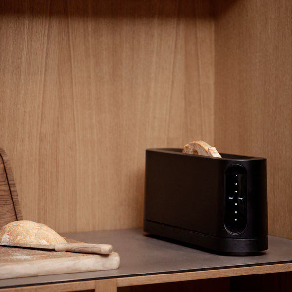 Ein schwarzer Toaster mit Brotscheiben neben einem Schneidebrett mit Brot auf einer hölzernen Küchenzeile.