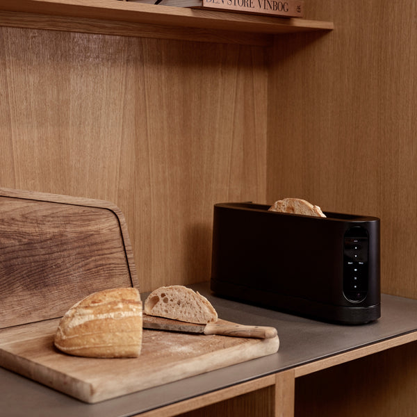 Aufgeschnittenes Brot und ein Messer auf einem Schneidebrett neben einem modernen schwarzen Toaster auf einer hölzernen Küchenzeile.