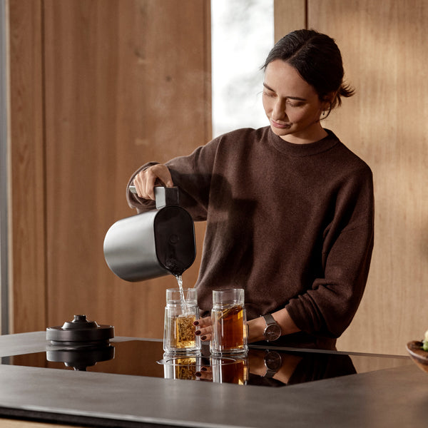 Frau gießt heißes Wasser aus einem Wasserkocher in einen Glasbecher mit Teebeuteln auf einer modernen Küchenzeile.