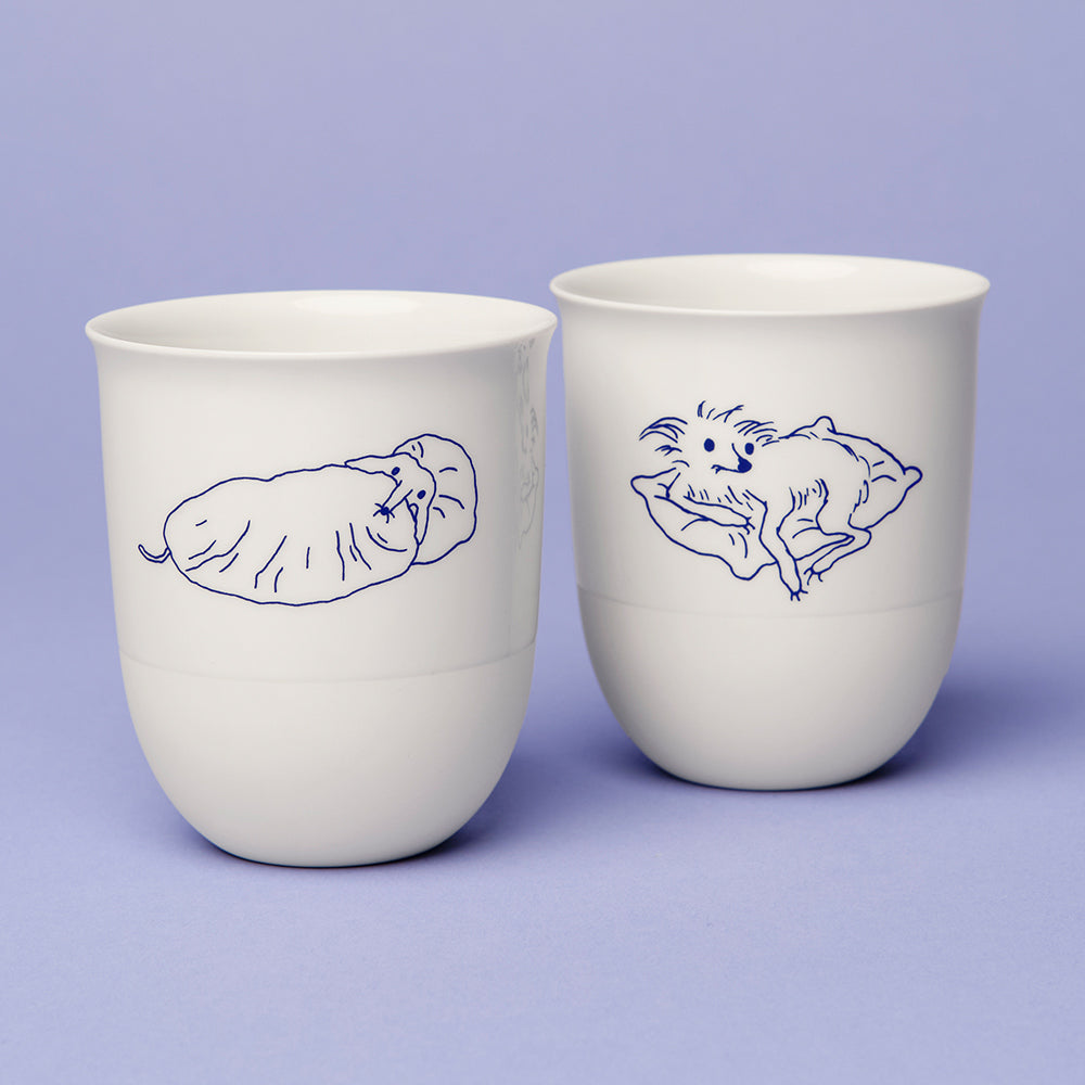 Auf einem hellvioletten Hintergrund sind zwei handgefertigte Porzellantassen mit Hundeillustrationen in blauer Linie auf weißem Grund abgebildet.