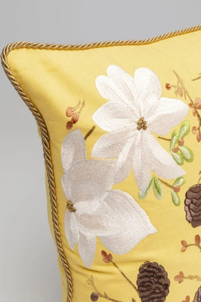 Ein gelbes Kissen mit aufgestickten weißen Blumen, braunen Tannenzapfen und grünen Blättern, das am Rand mit einem gedrehten Seil verziert ist.