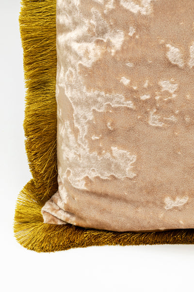Nahaufnahme eines beigen Samtkissens mit goldenem Fransenbesatz auf weißem Hintergrund.