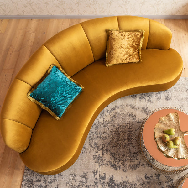 Geschwungenes senfgelbes Sofa mit teal- und goldfarbenen Kissen, daneben ein runder Couchtisch mit Birnen.