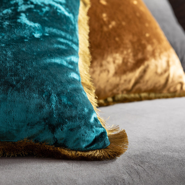 Nahaufnahme von blaugrünen und goldenen Samtkissen mit Fransen an den Rändern auf einer grauen Stoffoberfläche.