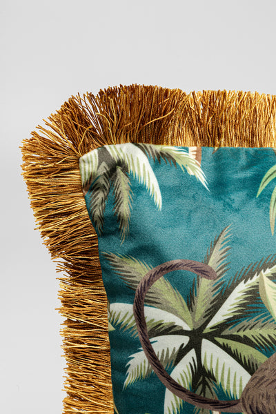 Nahaufnahme eines Kissens mit goldenen Fransen und tropischem Palmenblattdruck auf türkisfarbenem Stoff.