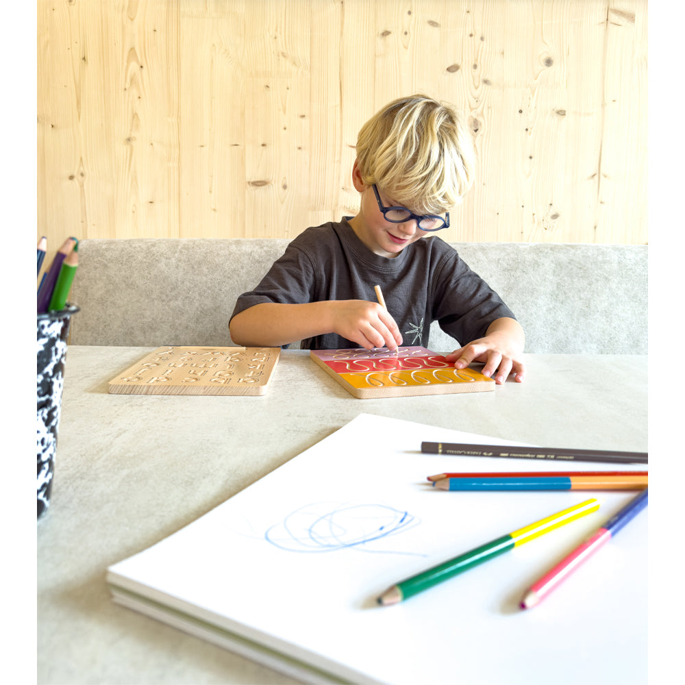 Ein kleiner Junge mit Brille zeichnet an einem Tisch Formen auf die Holztafel des Schreibtafel-Sets „Educate“, daneben liegen Buntstifte und Papier.
