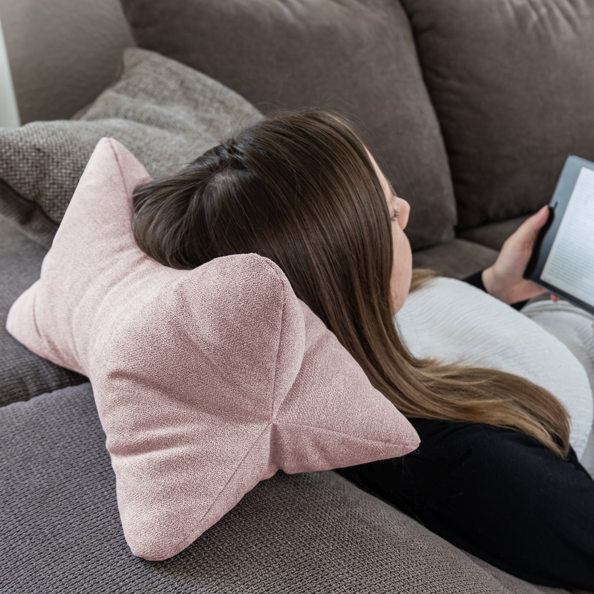 Eine Frau liegt auf einem Sofa, ruht mit dem Kopf auf einem Kissen von Leseknochen Cozy Home – Altrosa und liest auf einem Tablet.