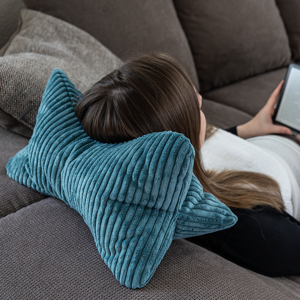 Eine Person lehnt sich auf einem Sofa zurück, legt ihren Kopf auf den Leseknochen Cozy Shara – Petrol als Nackenstütze und hält einen E-Reader für optimalen Lesekomfort.