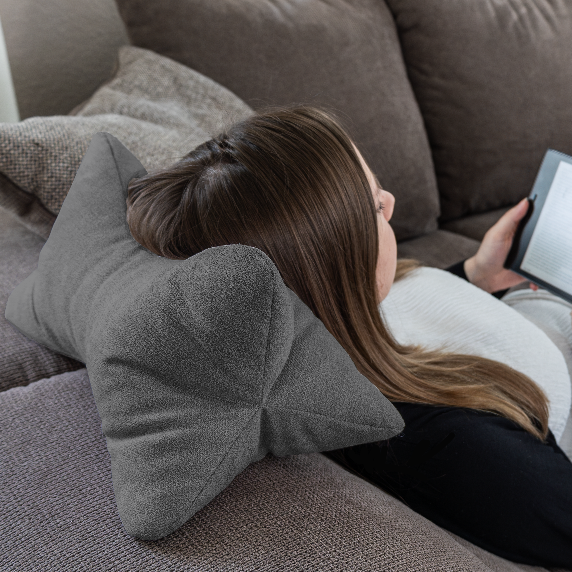 Eine Person liegt auf einer Couch und legt ihren Kopf auf den Leseknochen Cozy Home – Anthrazit, während sie für optimalen Komfort ein elektronisches Gerät liest.