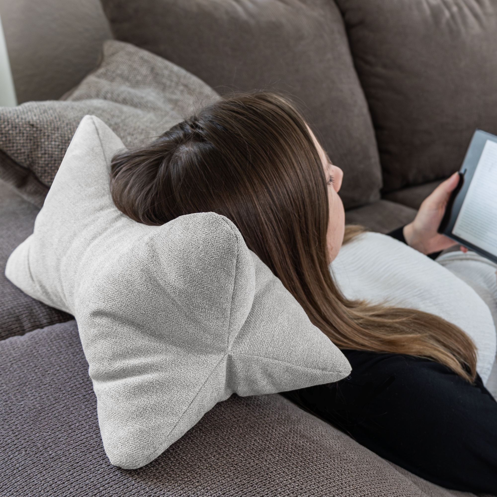 Eine Frau liegt auf einem grauen Sofa, ruht mit dem Kopf auf einem Leseknochen (Cozy Home – Grau) und liest auf einem elektronischen Gerät.
