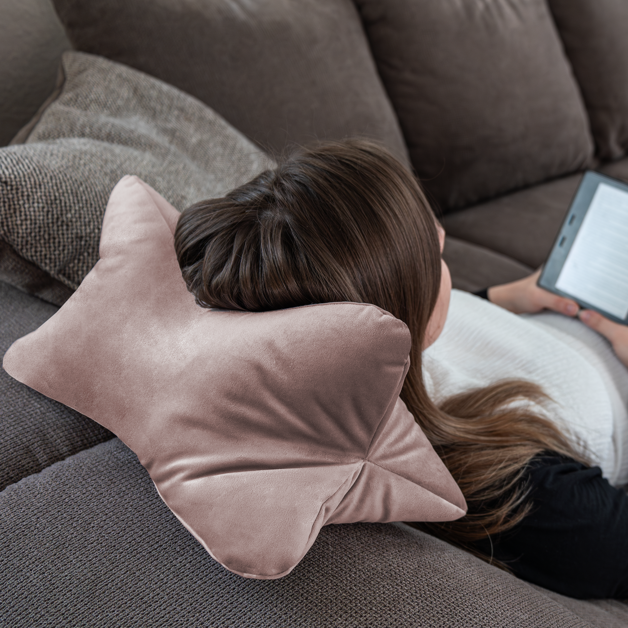 Eine Person mit langen braunen Haaren entspannt auf einer Couch, nutzt das Leseknochen Cozy Samt - Rose Kissen als Nackenstütze und hält einen E-Reader oder ein Tablet in der Hand und genießt optimalen Komfort.