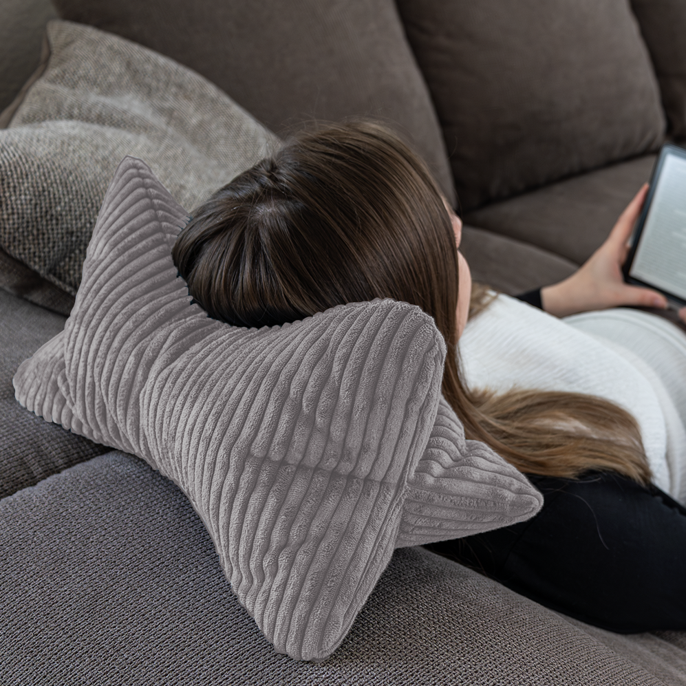 Eine Person liegt auf einem Sofa, ruht mit dem Kopf auf dem Leseknochen Cozy Shara – Anthrazit, einem ergonomisch geformten Nackenstützkissen in Knochenform, und hält einen E-Reader in der Hand.
