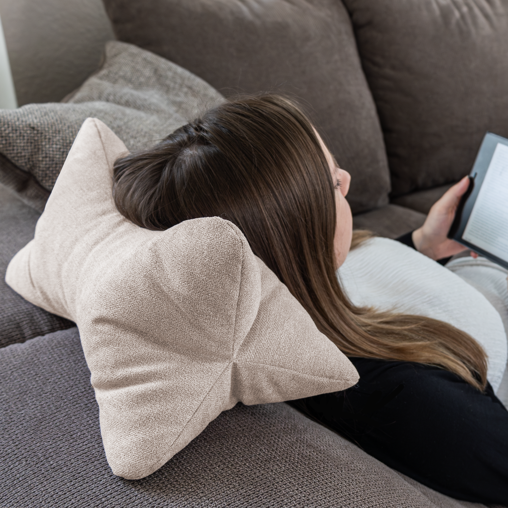 Eine Frau liegt auf einem Sofa, ihr Kopf ruht auf einem Leseknochen Cozy Home - Beige-Kissen, während sie auf einem E-Reader liest.