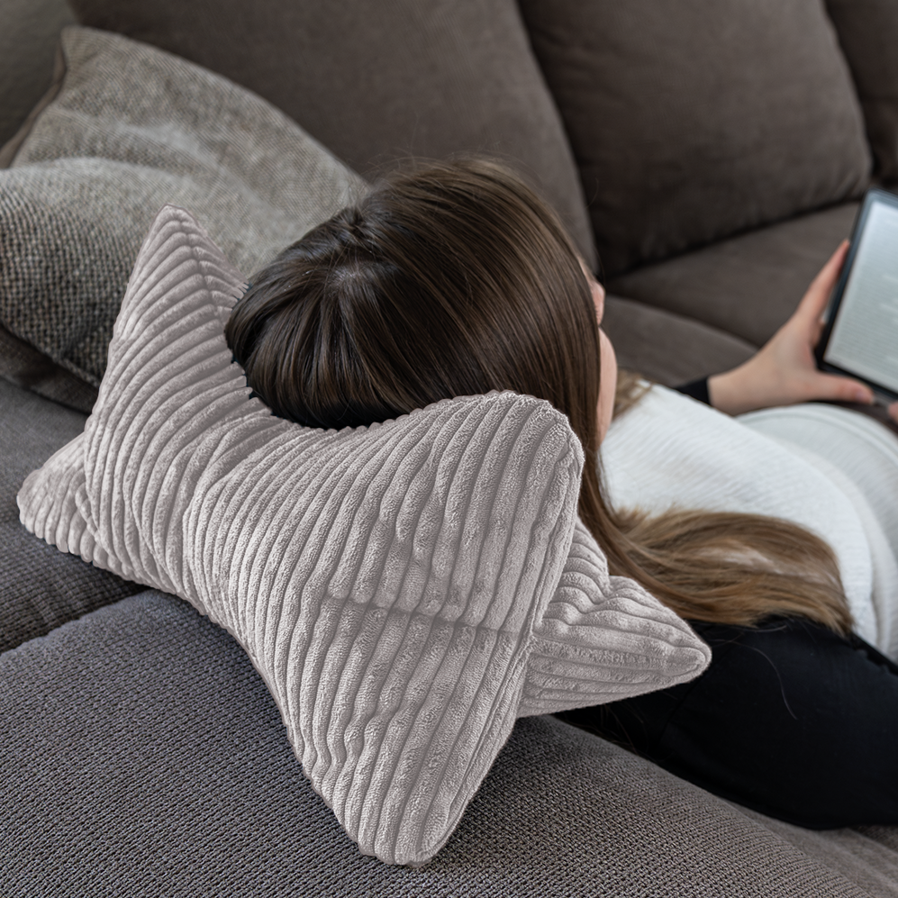 Eine Person lehnt sich auf einem Sofa zurück, nutzt den Leseknochen Cozy Shara – Grau als stützende Nackenstütze, hält einen E-Reader und genießt Komfort und eine gesunde Körperhaltung.