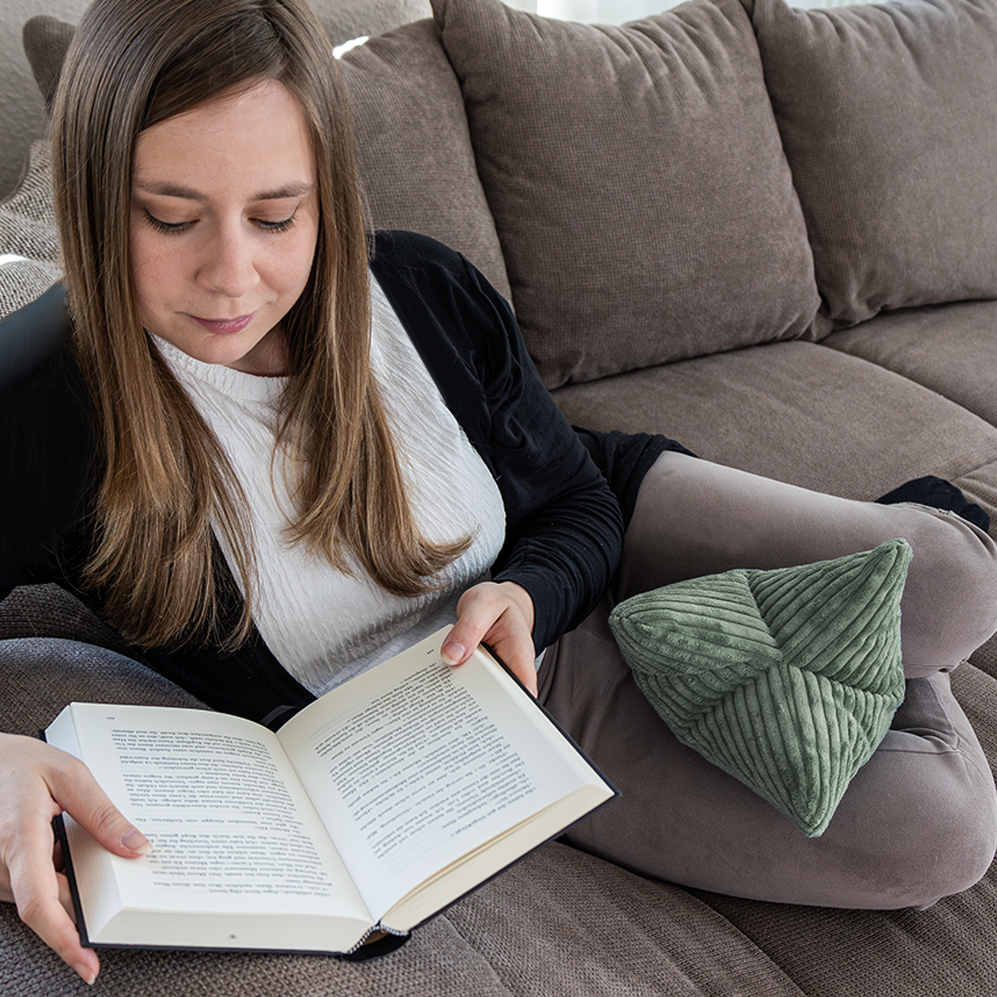Eine Frau sitzt auf einem grauen Sofa und liest. Der Leseknochen Cozy Shara – Salbei dient als Nackenstütze für optimalen Komfort, während ein grünes strukturiertes Kissen auf ihren übereinandergeschlagenen Beinen ruht.