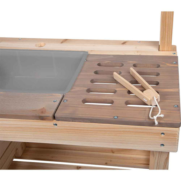 Eine Nahaufnahme eines hölzernen Outdoor-Küchenspielsets mit einer grauen Plastikspüle, einer geschlitzten Holzfläche und einer Holzzange darauf.
