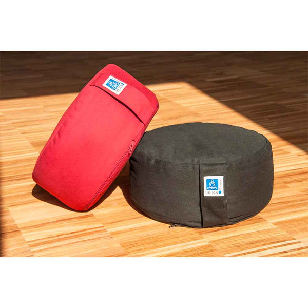 Ein rotes und ein schwarzes Meditationskissen liegen auf einem Holzboden im Sonnenlicht.