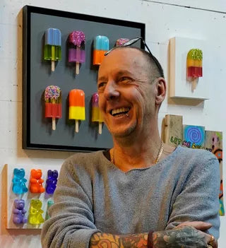 Der Künstler Michael Pfannschmidt steht lächelnd vor einer bunten Eiszapfen- und Gummibärchenkunst an der Wand.