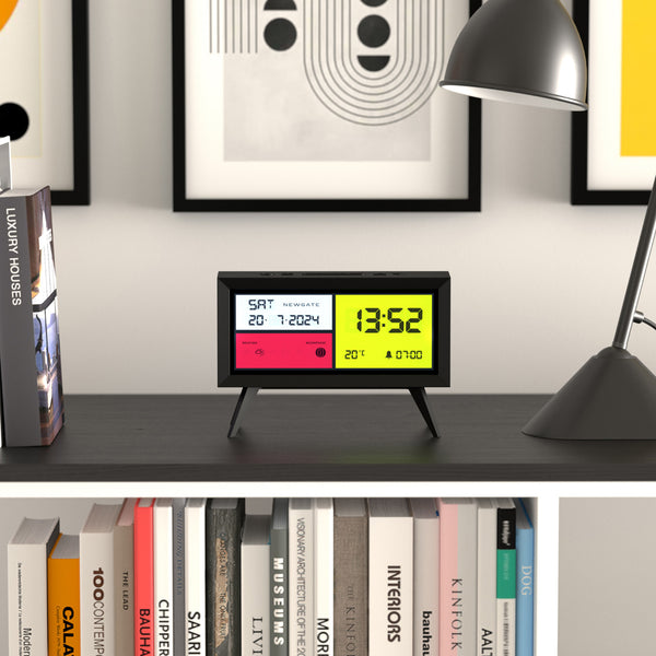 Moderne Digitaluhr auf einem Schreibtisch mit Büchern, Lampe und gerahmter Kunst im Hintergrund.