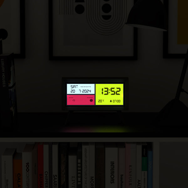 Eine Digitaluhr auf einem Regal zeigt neben Büchern und gerahmter Kunst das Datum, die Uhrzeit, die Temperatur und den Alarm an.